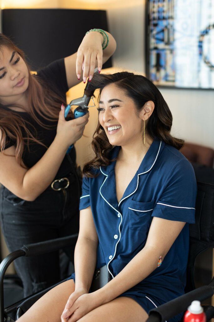 A stylist uses a curling iron to curl a smiling woman's hair, who is seated and wearing blue pajamas.