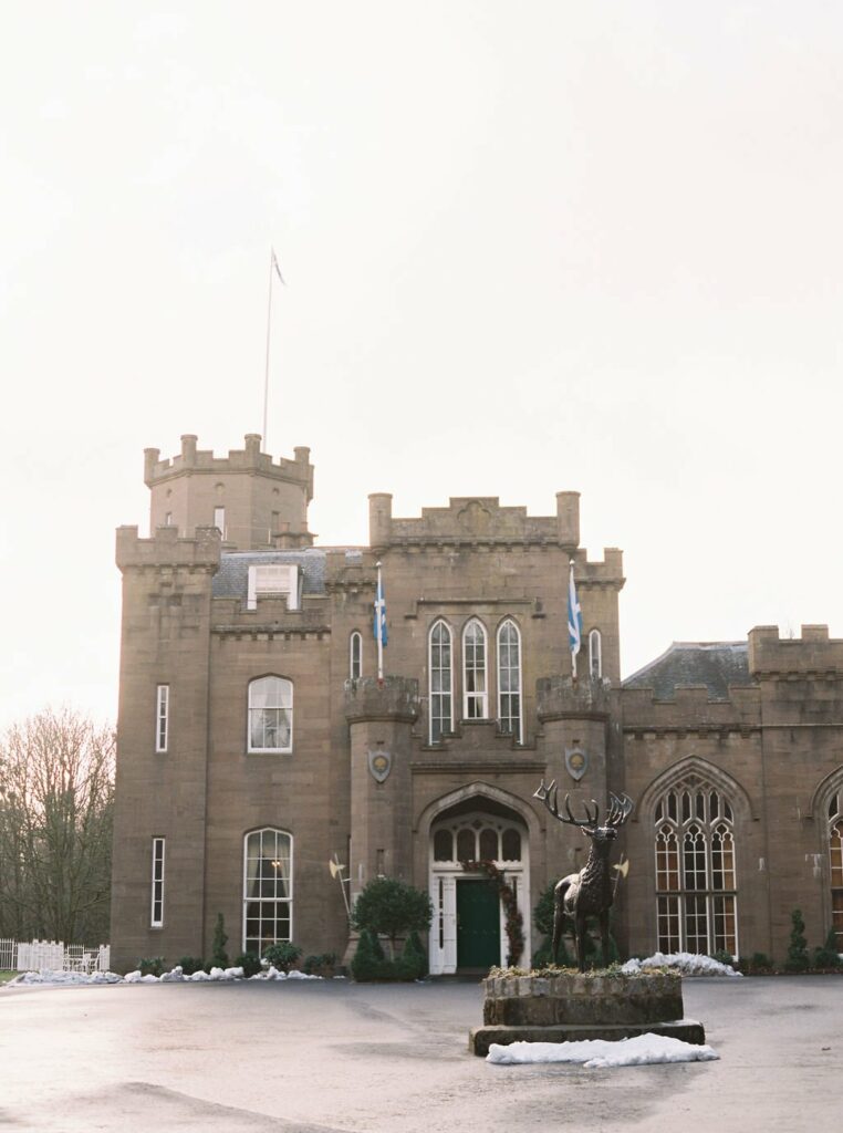 A large, castle-like building with two Scottish flags, arched windows, and a statue of a stag in front. There is a light covering of snow on the ground.