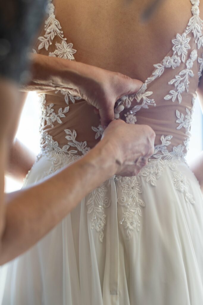 Close-up of a person fastening the back of a white, lace-embroidered wedding dress for another person.