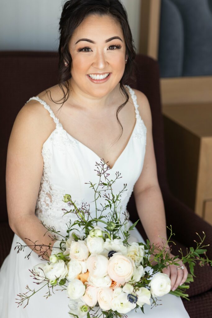 A woman in a white wedding dress holds a bouquet of white and pale peach flowers while sitting on a dark chair and smiling.