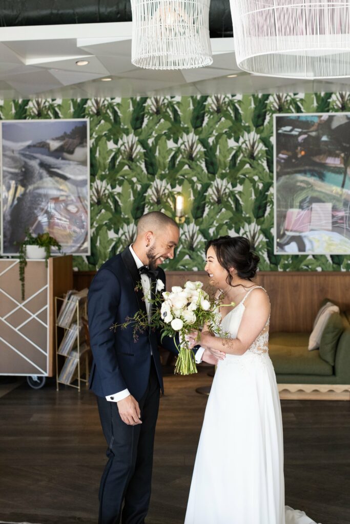 A bride and groom exchange smiles while holding bouquets in a room with green, patterned wallpaper and framed art.
