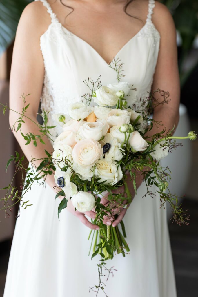 A woman in a white dress holds a bouquet of white roses and greenery. Her face is out of the frame.