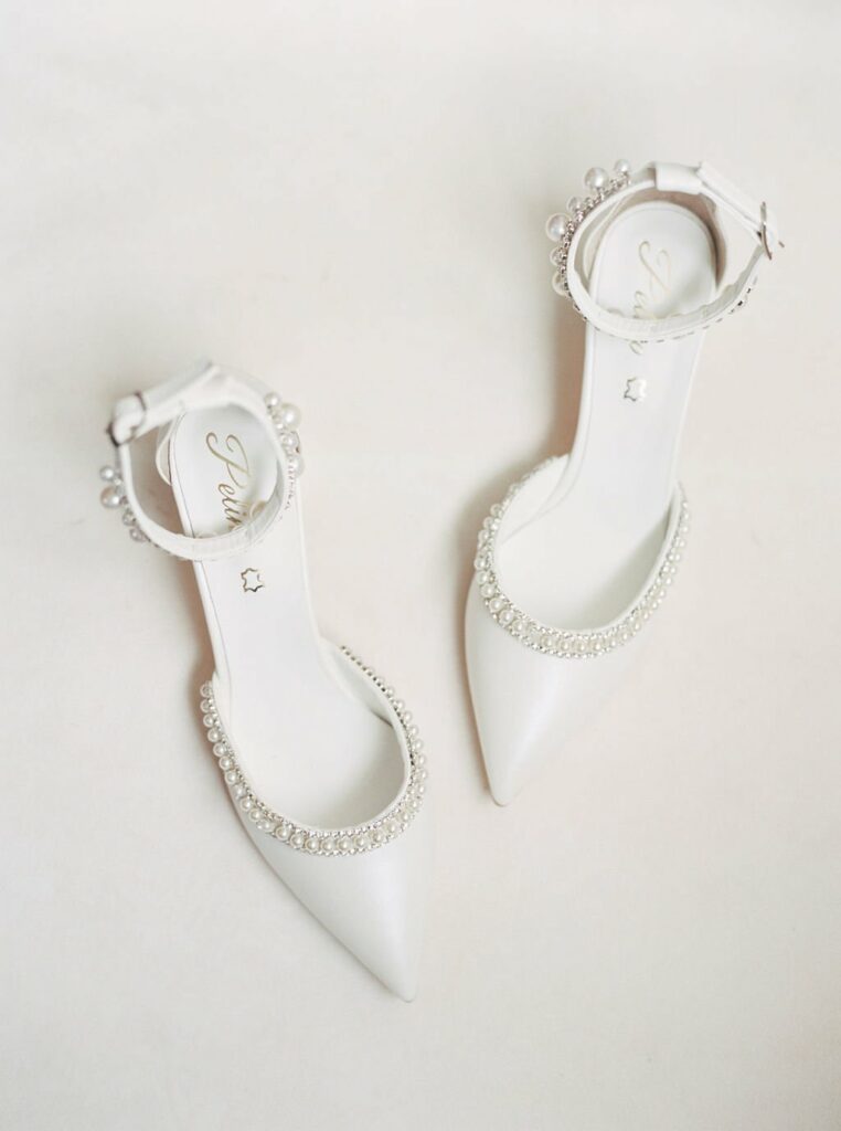 A pair of white high-heeled shoes with ankle straps, decorated with pearls and rhinestones, placed on a light background.