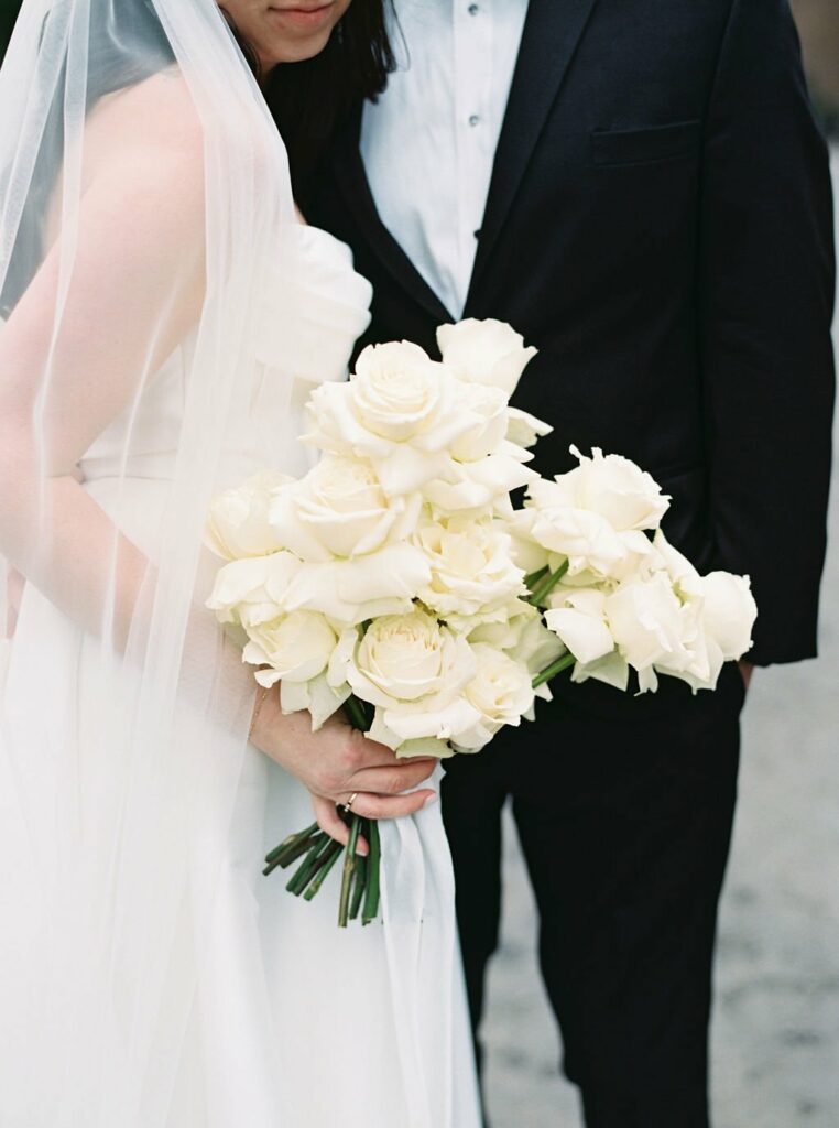 Bride in white dress and veil holding a bouquet of white roses, standing next to a person in a black suit. Faces are not visible.