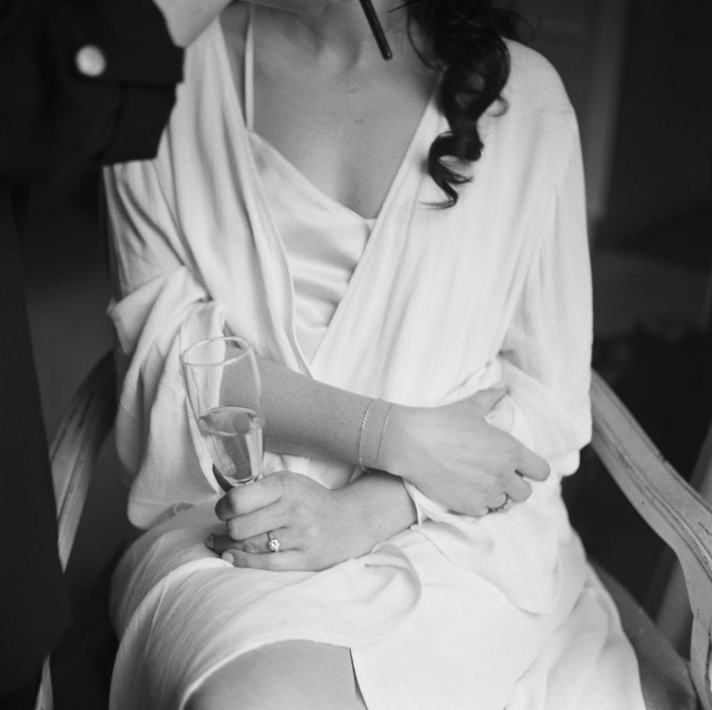 A person in a white robe holds a champagne flute while sitting on a chair, with another person's hand applying makeup to their face.