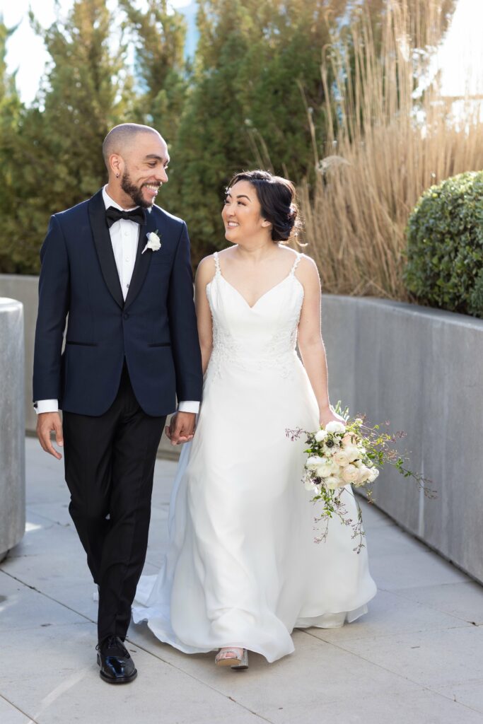 A couple dressed in wedding attire, the groom in a dark suit and bow tie and the bride in a white gown holding a bouquet, walk hand in hand outdoors, smiling at each other.