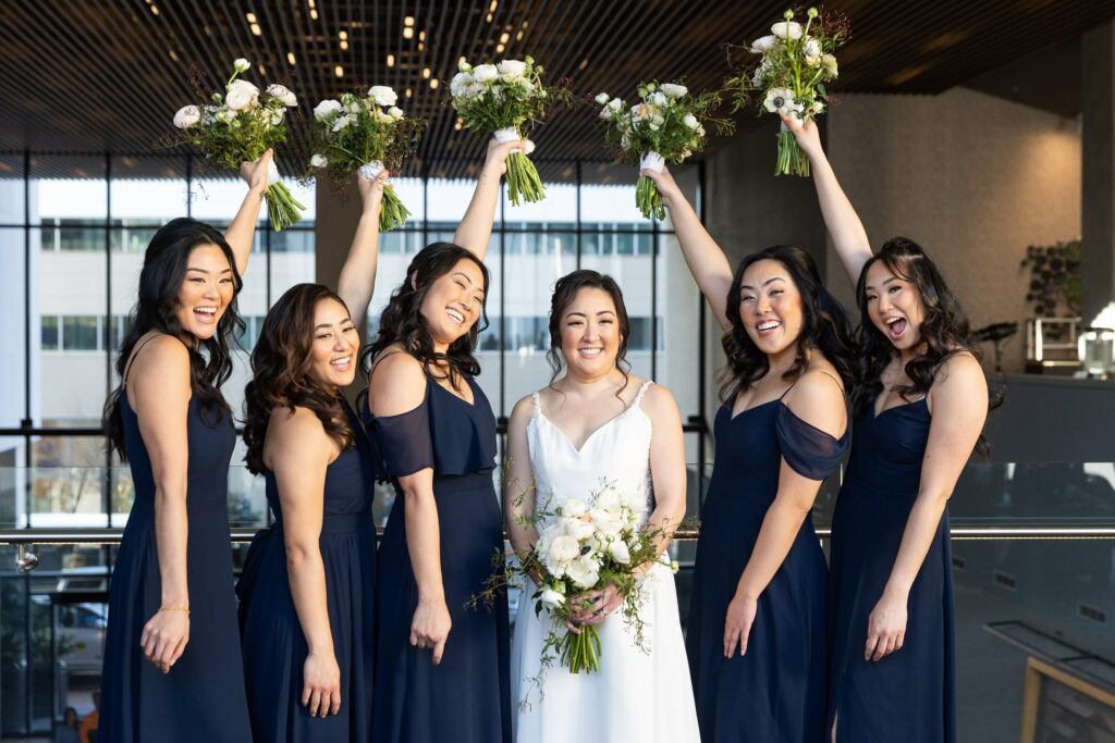 A bride stands smiling in a white gown surrounded by five bridesmaids in matching navy dresses, raising their floral bouquets in celebration.