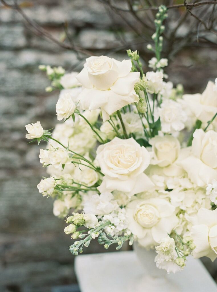 A floral arrangement featuring white roses and other white flowers in a white vase, set against a blurred stone background.