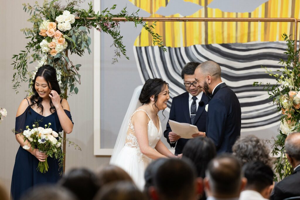 A bride and groom hold hands and smile at each other during their wedding ceremony, with a bridesmaid holding a bouquet and an officiant reading from a paper. Floral decorations surround the scene.