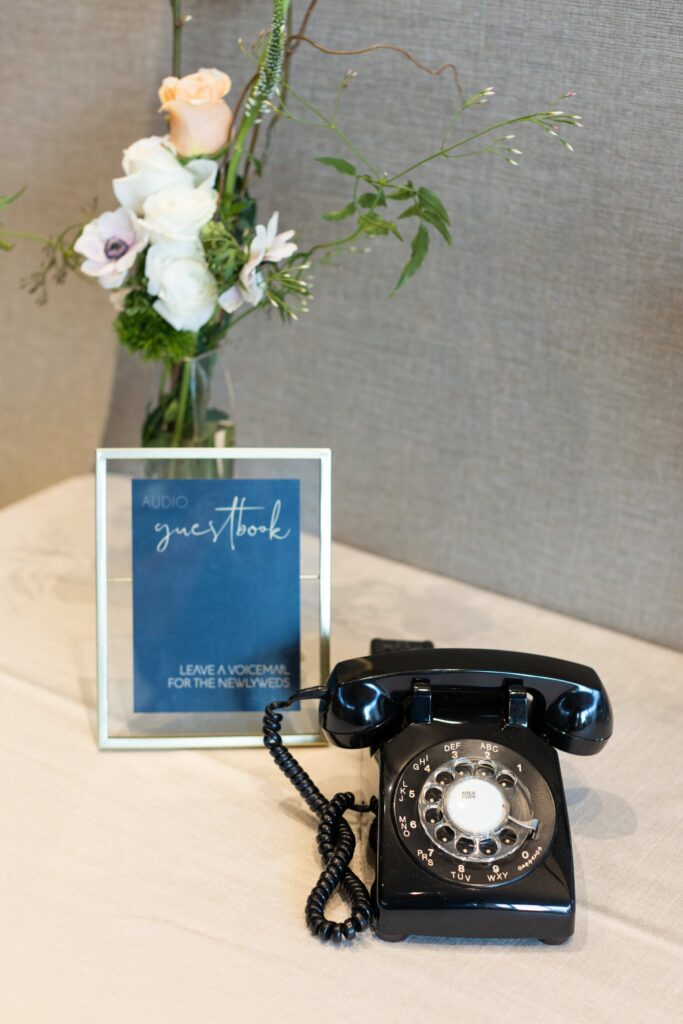 A vintage black rotary phone is placed on a table beside a blue sign that reads "Audio Guestbook: Leave a Voicemail for the Newlyweds," with a floral arrangement in the background.