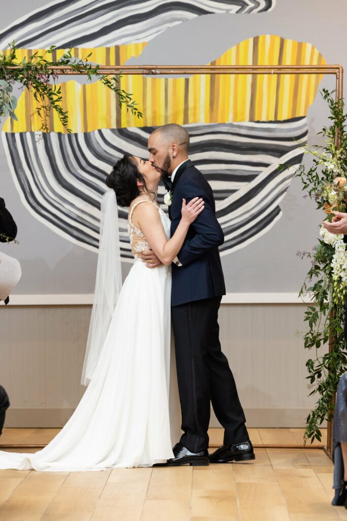 A couple in a wedding ceremony is kissing under a decorated arch. The bride is wearing a white dress and veil, and the groom is in a dark suit. A piece of modern art is visible in the background.