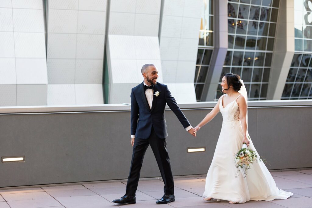 A bride and groom in formal attire hold hands and walk together on an outdoor terrace with a modern building in the background.