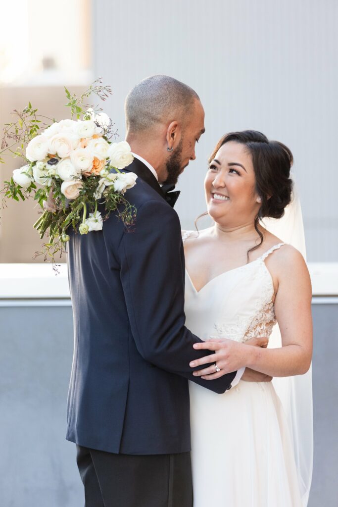 A bride and groom are standing close together. The groom is holding a bouquet of white and peach flowers, and they are looking at each other with smiles.