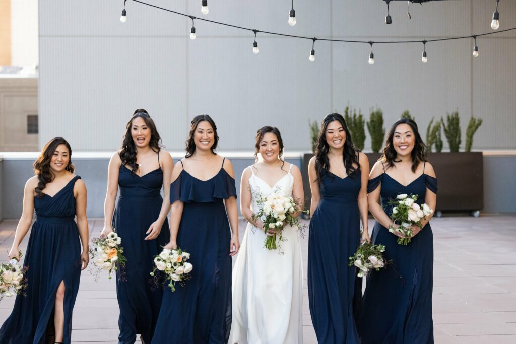 A bride in a white gown stands with five bridesmaids in navy blue dresses, each holding a bouquet. They are outdoors under string lights.