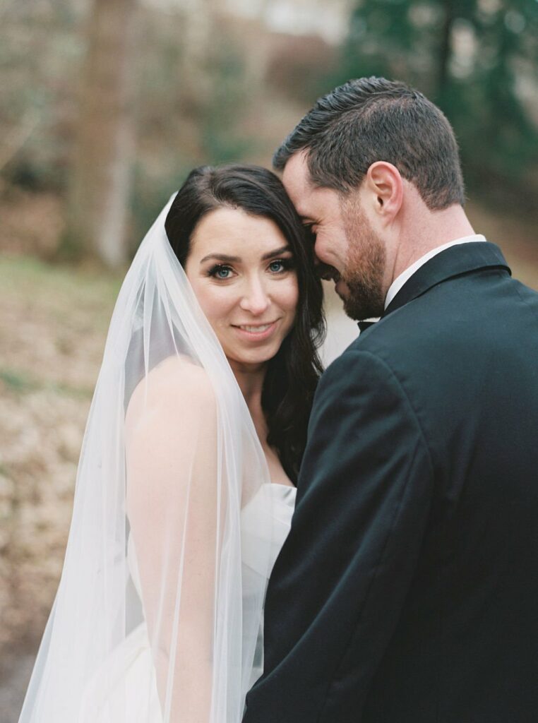 A bride in a white dress with a veil smiles while holding hands with a groom in a black suit outdoors.