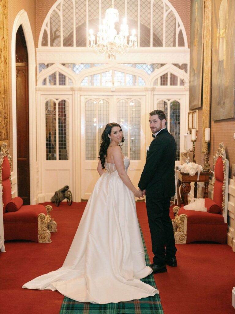A bride and groom stand holding hands in an ornate room with red carpet, white trim, and large windows. The bride wears a white gown with a long train, and the groom is in a black suit.