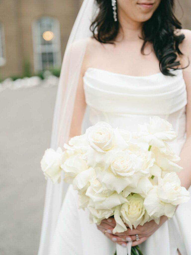 A bride in a strapless white dress holding a bouquet of white roses, with a brick building in the background.