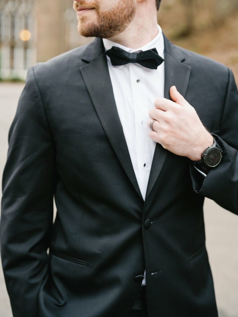 Man in a black tuxedo with a bow tie adjusting his collar, wearing a wristwatch and standing outdoors.
