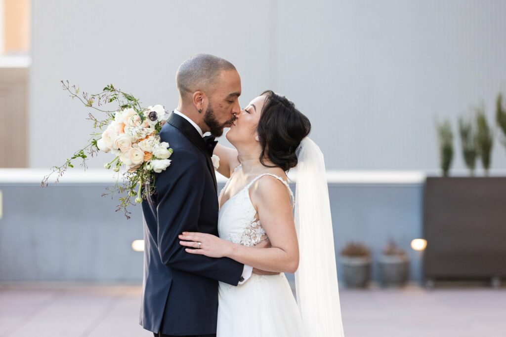 A bride and groom share a kiss on their wedding day. The groom is holding a bouquet of flowers, and the bride is wearing a white wedding dress with a veil.