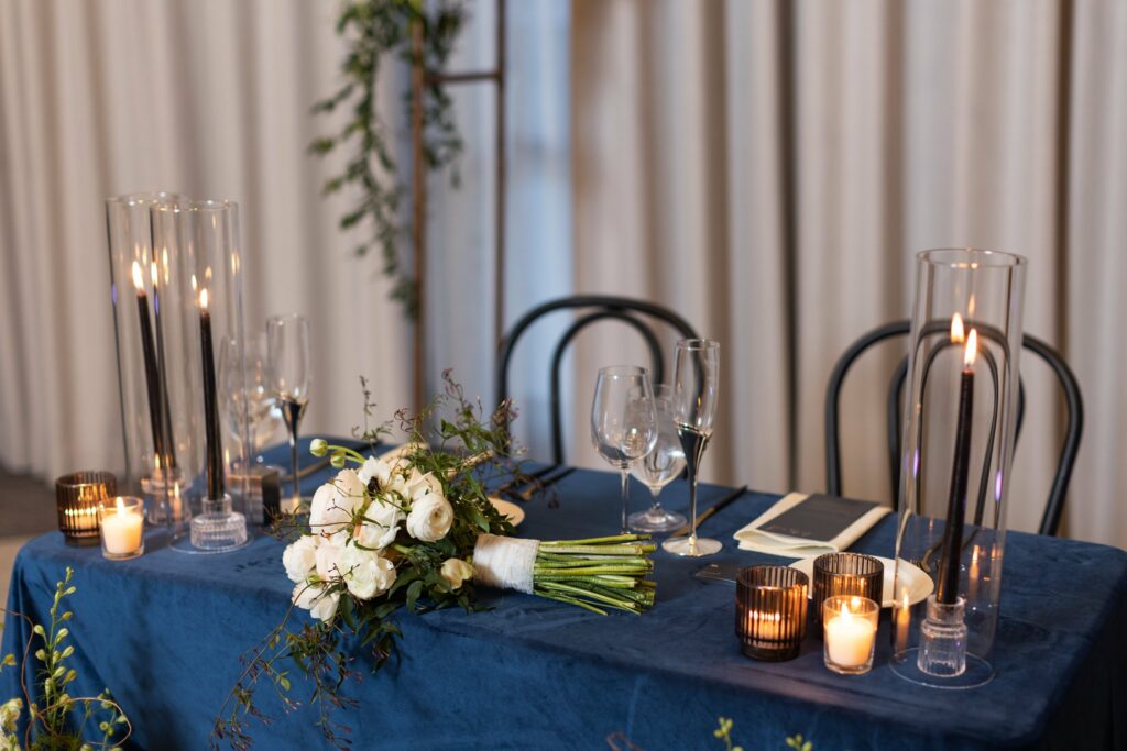 A table with a blue tablecloth is set with floral arrangements, wine glasses, candles, and a bouquet of white flowers.