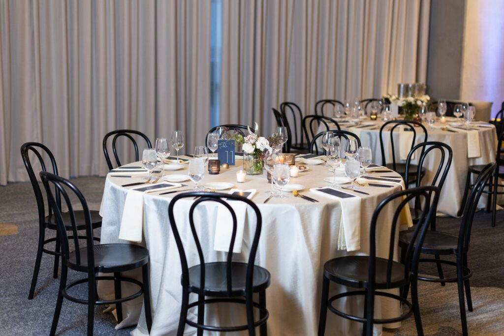 A dining setup with round tables covered in white tablecloths, black chairs, glassware, and floral centerpieces. Candlelights are placed on the tables, and curtains are drawn in the background.