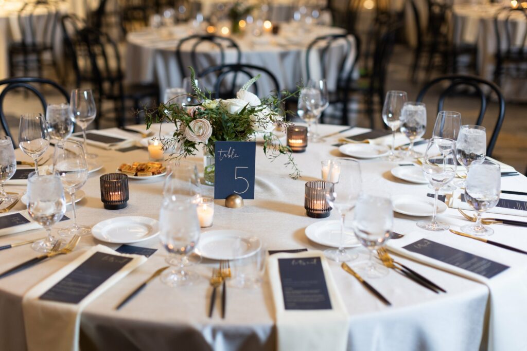 A round table set for a formal event, labeled "Table 5" with floral centerpieces, glassware, utensils, menus, bread plates, and water glasses on white tablecloths. Background shows similar tables.