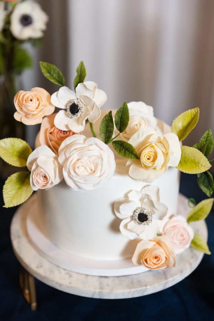A white cake decorated with realistic-looking sugar flowers, including roses and anemones, and green leaves, displayed on a marble cake stand.
