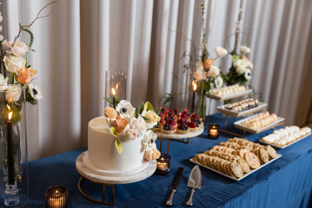 A dessert table with a white floral cake, various pastries, cookies, candles, and floral arrangements on a blue tablecloth.