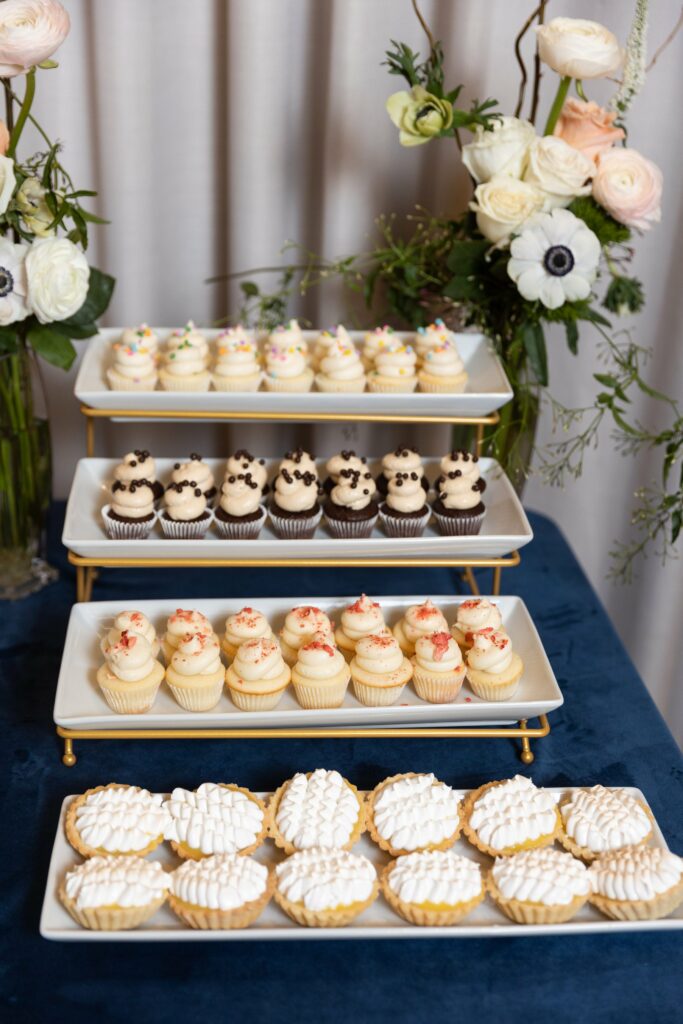A table displays a three-tiered stand of assorted cupcakes and a separate tray of small tart desserts, surrounded by floral arrangements.