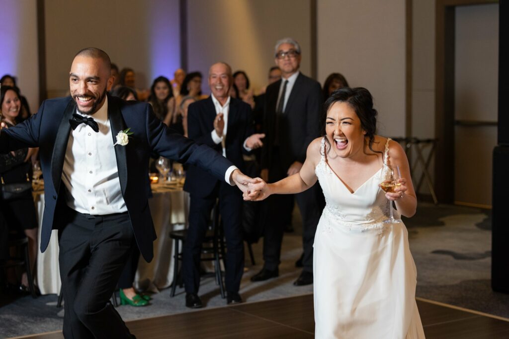 A bride and groom hold hands and laugh joyfully while walking on a dance floor, with smiling guests applauding in the background during a wedding reception.