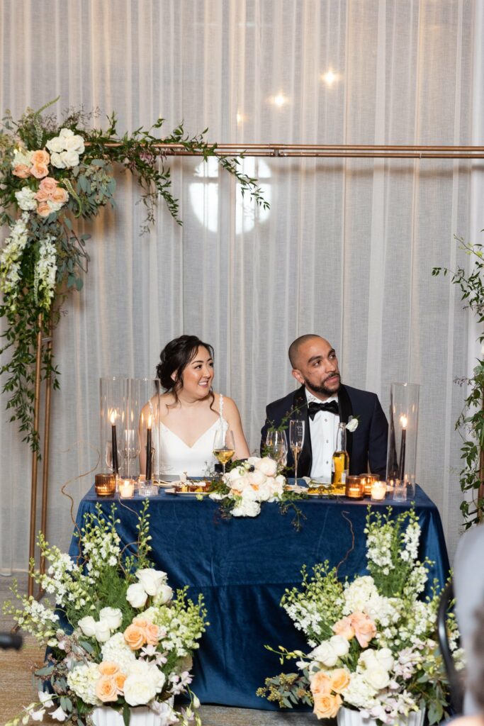 A couple in formal attire sits at a decorated table with flower arrangements and candles. They both look to the side while seated in a reception-like setting.