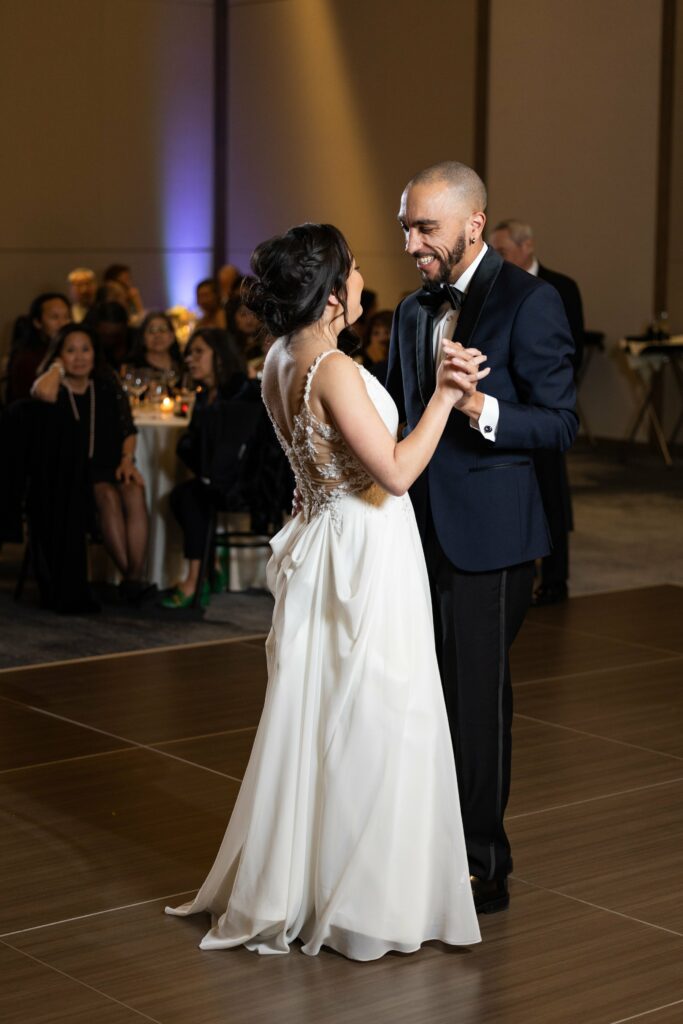 A bride and groom share a dance at their wedding reception, surrounded by seated guests. The bride wears a white dress with a detailed back, and the groom is in a dark suit.