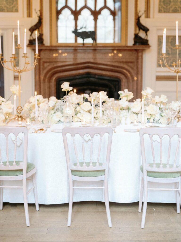 A white tablecloth-covered dining table with white floral arrangements, candles, and three chairs is set in front of a decorative fireplace with ornate gold accents.