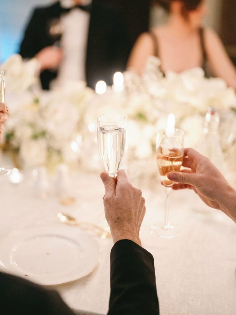 Closeup of two hands clinking champagne glasses at a formal event, with blurred background featuring white floral arrangements and other attendees.