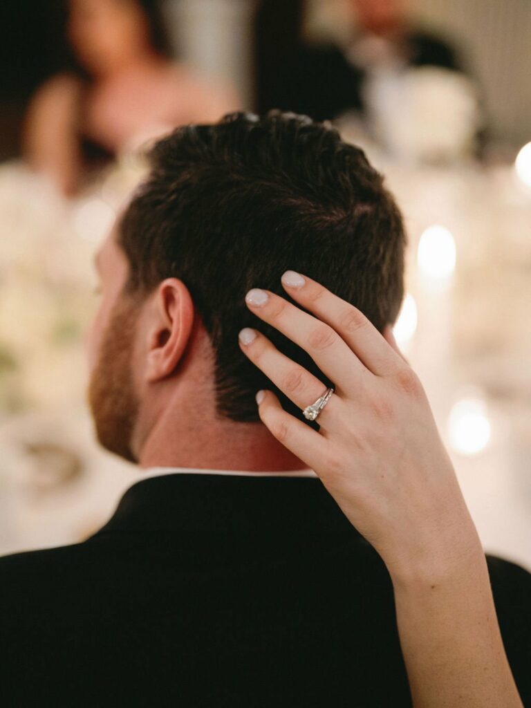 A woman's hand with a large engagement ring rests on the back of a man's head as they sit at a table decorated with white flowers and lit candles.