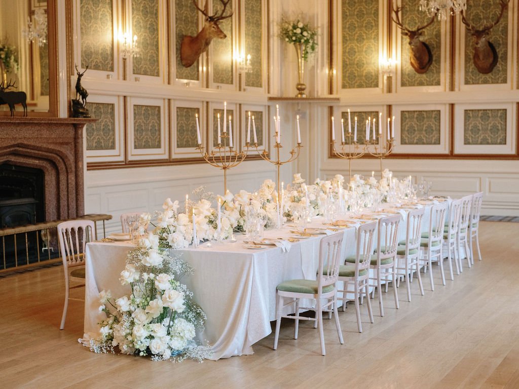 A long dining table set elegantly with white floral arrangements and gold candlesticks in an opulent room with ornate wall paneling, chandeliers, and mounted deer heads.
