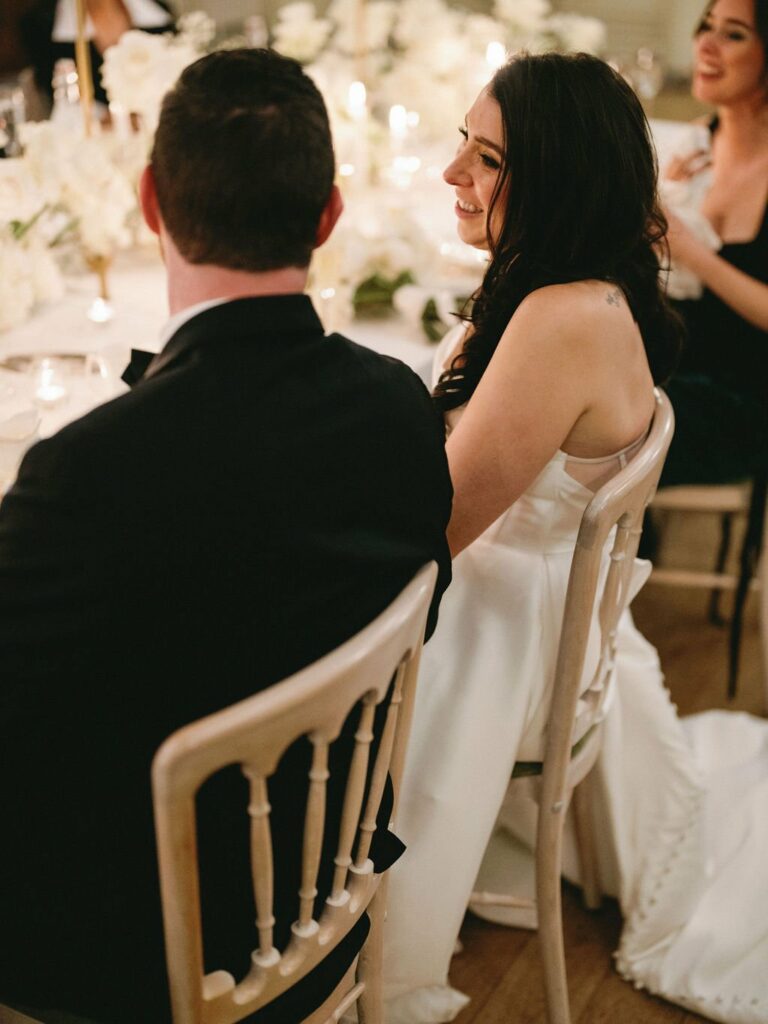 A bride in a white dress and a groom in a black suit sit at a decorated table during a wedding reception with a guest in the background.