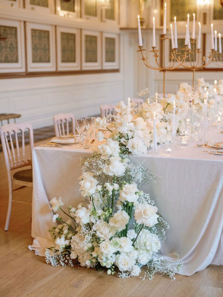 A long table decorated with a white tablecloth is set with white floral arrangements, candles, and elegant tableware in a sophisticated indoor setting.