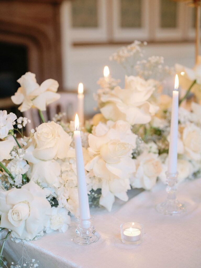 A close-up of white roses and other flowers arranged with three lit white taper candles and a single tealight candle on a table.