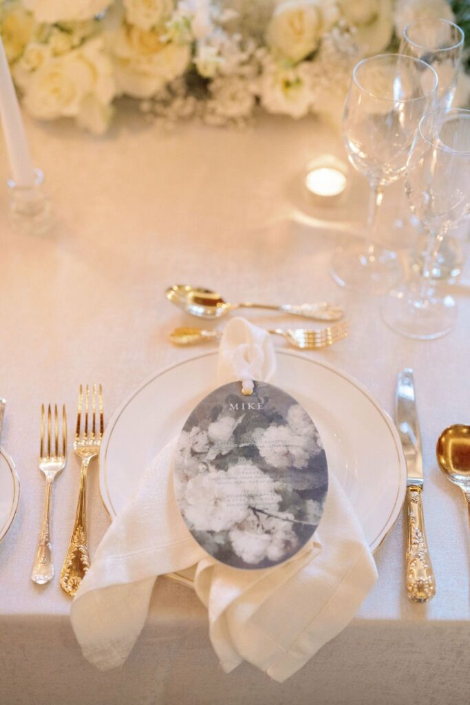 Table setting with white dinnerware, gold cutlery, a napkin with a floral menu card, and floral centerpiece. Glassware, a candle, and a white tablecloth complete the elegant arrangement.