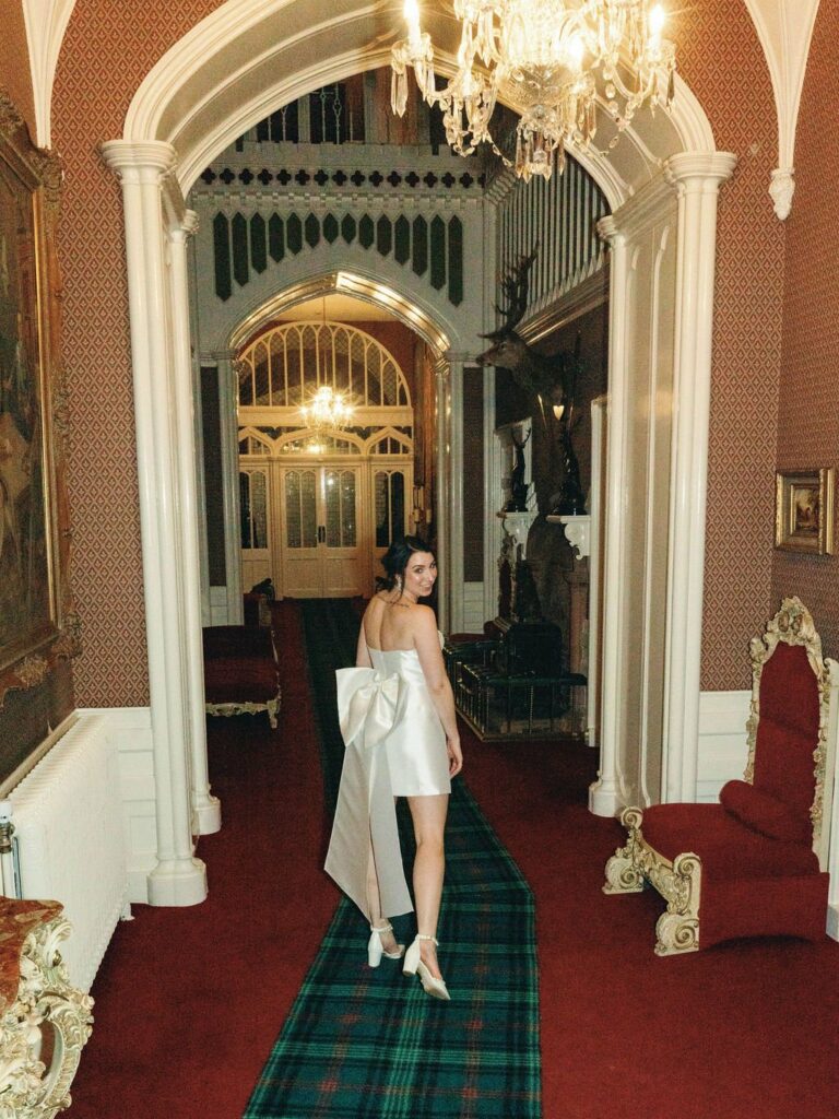 A woman in a white dress with a large bow walks down a grand, ornate hallway with chandeliers and red carpeting.