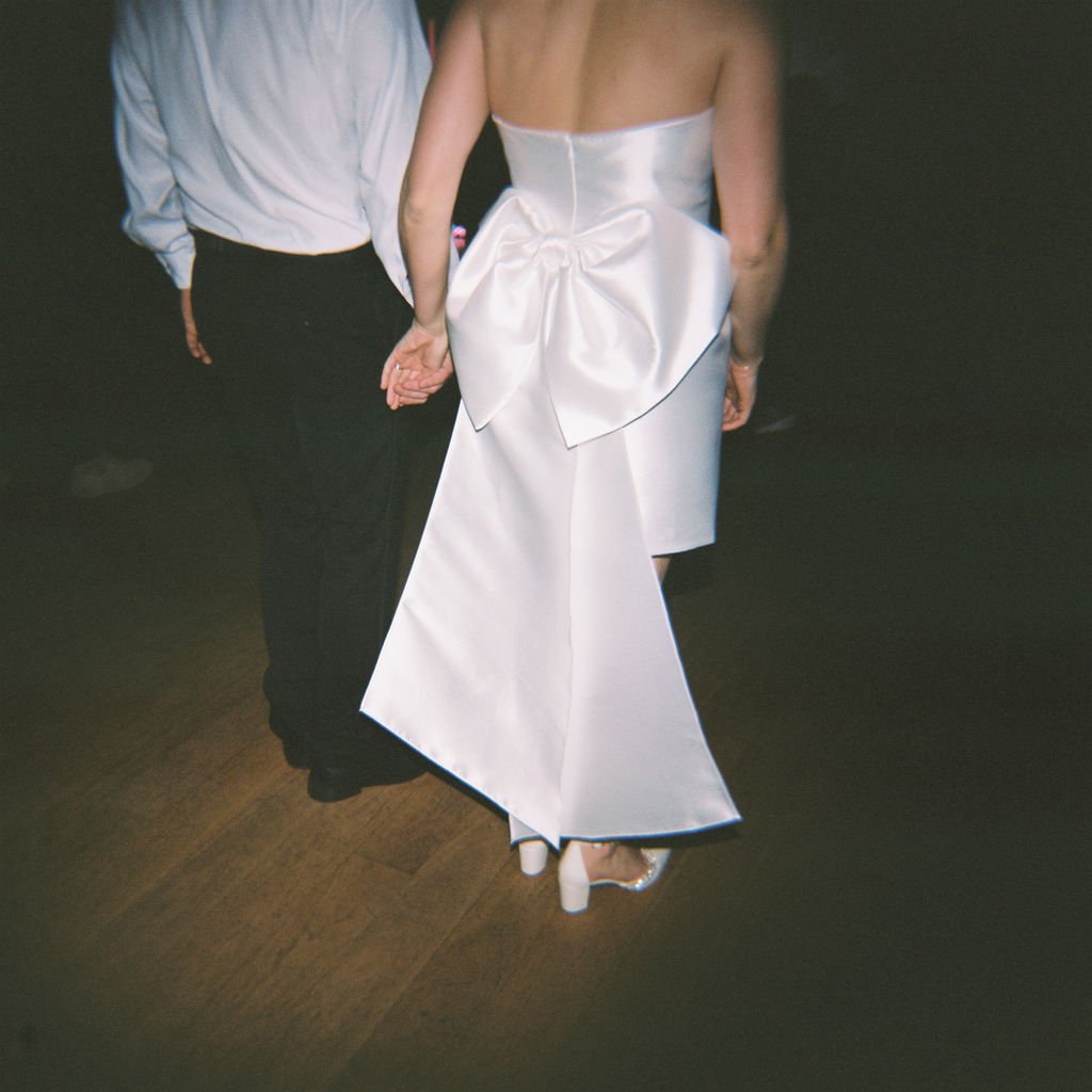 A couple stands holding hands; the woman wears a white dress with a large bow on the back, and the man wears black pants and a white shirt.