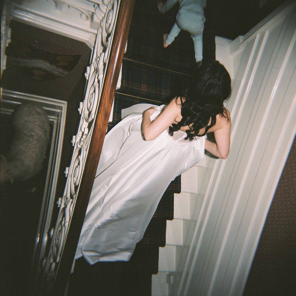 A person in a white dress is walking up a staircase, with another person, partially visible, ahead of them. The scene includes intricate railing and carpeting on the stairs.
