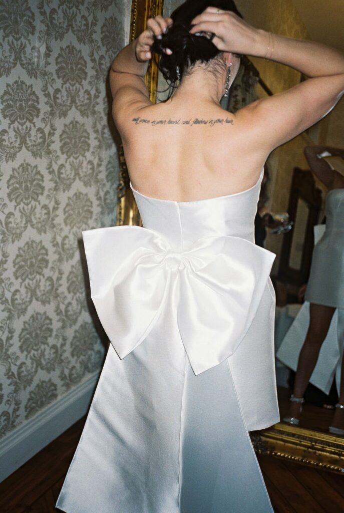 A person in a white strapless dress with a large bow on the back is adjusting their hair in front of a mirror. An upper back tattoo is partially visible. They stand in a room with patterned wallpaper.