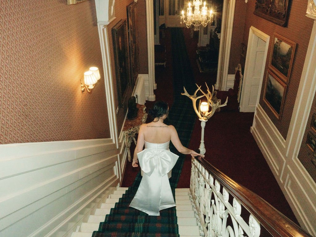 A woman wearing a white dress with a large bow descends a carpeted staircase in a dimly lit, ornate interior with framed pictures on the walls and chandeliers.