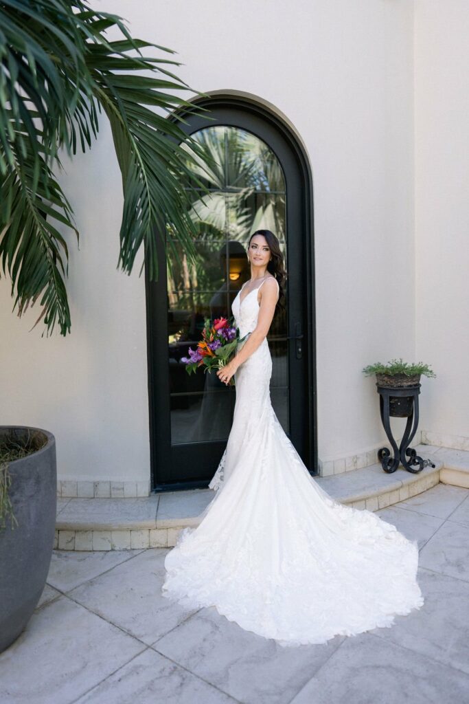 A woman in a white wedding dress holds a colorful bouquet, standing in front of a black arched door framed by greenery.