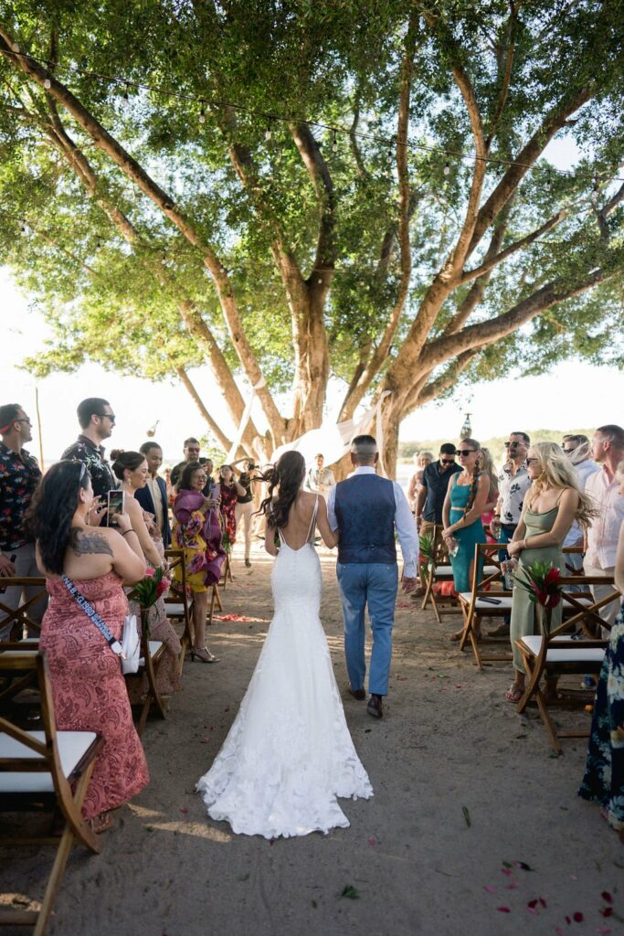 A bride and groom walk down an outdoor aisle under a large tree, surrounded by standing guests dressed in semi-formal attire. The bride wears a white dress and the groom a blue vest with grey pants.