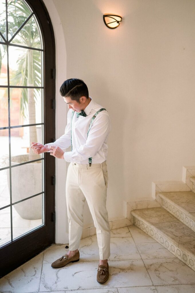 A man dressed in light-colored trousers, a white shirt, and suspenders adjusts his cuffs while standing near a doorway inside a stylishly decorated room with tiled floors.