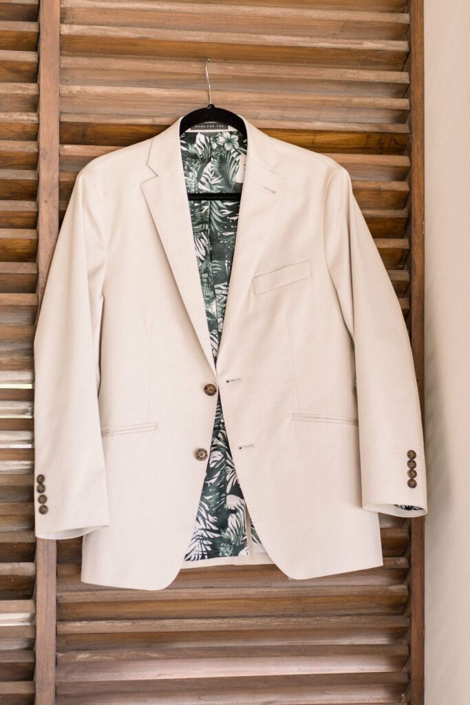 A beige blazer with brown buttons hangs on a wooden louvered shutter. The blazer's interior features a tropical leaf pattern.