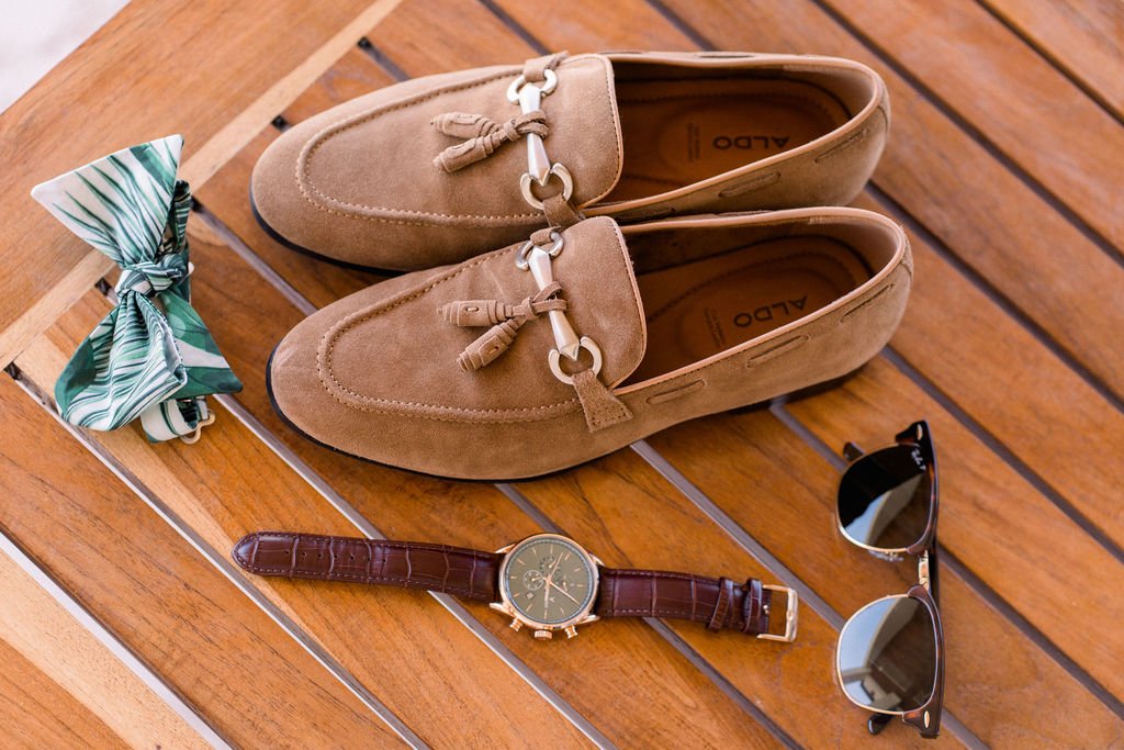 Tan suede loafers, a green and white patterned bow tie, a brown leather wristwatch, and aviator sunglasses are neatly arranged on a wooden surface.